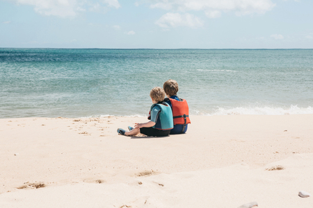 Two brothers or a twin or a friend in life jackets are sitting on the beach in the sand and looking at the sea or the ocean and dreaming. Pastime together. Childrens friendship or family.の写真素材