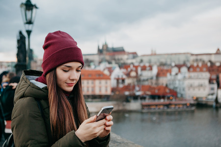 A young beautiful tourist girl stands on the Charles Bridge in Prague in the Czech Republic and uses a mobile phone to call or view a map or mobile application.の写真素材
