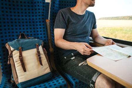 A tourist or a guy or a student with a backpack moves by train. He holds a map in his hands and looks out the window while the train is moving.の写真素材