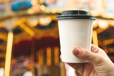 A young girl holds a disposable glass with coffee or another hot drink in the cold season. Carousel or merry-go-round and celebration in the background.の写真素材