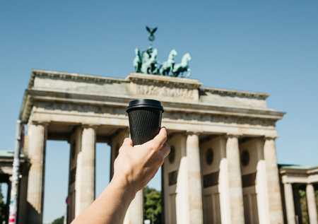 A person holds a disposable cup with coffee or another drink on the background of the Brandenburg...