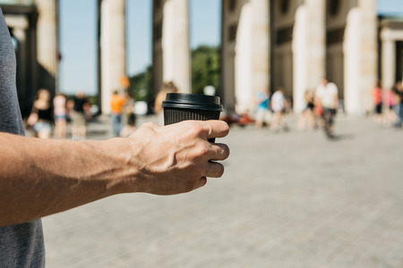 Close-up. A person holds a disposable cup with coffee or another drink while walking or sightseeing in Berlin, Germany. Ahead is the Brandenburg Gate and blurred, unrecognized people.の写真素材