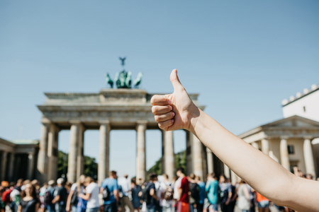 A person shows a sign with a finger upwards meaning everything is okay or confirms against the background of the Brandenburg Gate and blurred unrecognizable people in Berlin in Germany.の写真素材