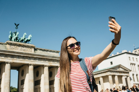 Young beautiful positive girl makes selfie against the background of the Brandenburg Gate in Berlin in Germany or takes pictures of sights.の写真素材