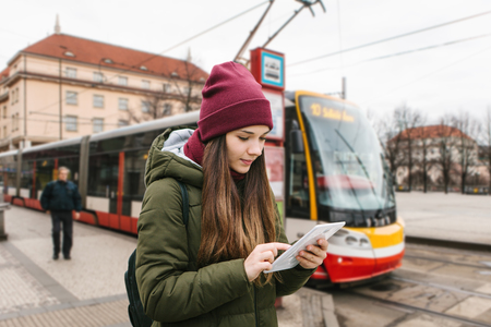A girl or a tourist got off the tram stop from a tram and looks at a tablet city map or a mobile application. She uses the internet or she is online. In the background unrecognizable person and tramの写真素材