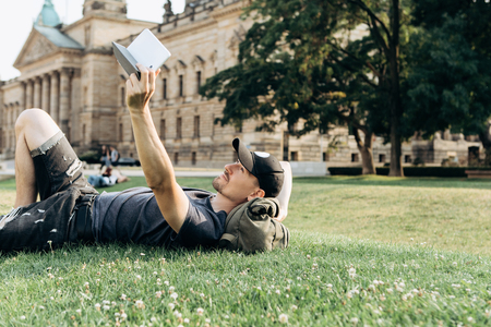 A young guy with a backpack or a student is lying on the grass during a break or is resting and using a tablet. He is watching a video or reading an e-book or something else.の写真素材