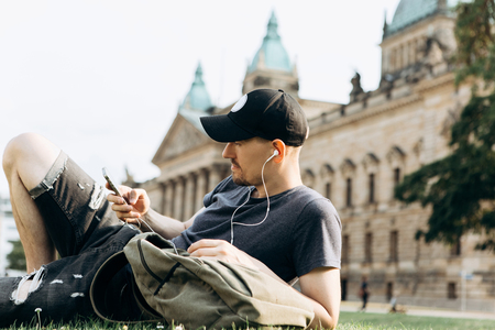 A young guy with a backpack or a student lies on the grass and listens to music or a podcast and enjoys.の写真素材