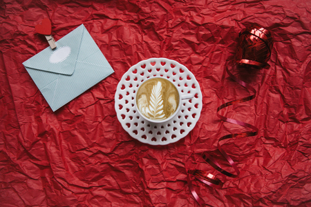 Delicious cappuccino coffee on a red background. Near an envelope with a letter or a greeting and a ribbon. Celebration of Valentines Day or another love event.の写真素材