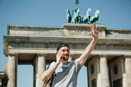 A tourist or a guy with a backpack is talking on a mobile phone and is greeting someone with his hand. In the background is the Brandenburg Gate in Berlin in Germany.の写真素材
