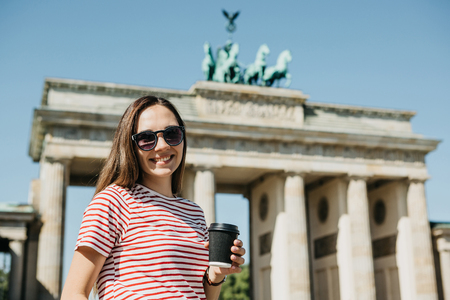 Portrait of a young beautiful positive smiling girl who holds in her hand a coffee or other drink on the background of the Brandenburg Gate in Berlin in Germany.の写真素材