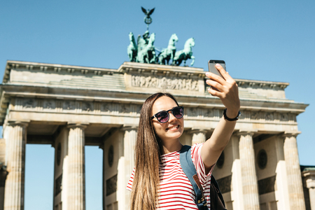 Young beautiful positive girl makes selfie against the background of the Brandenburg Gate in Berlin in Germany or takes pictures of sights.の写真素材