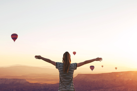 A girl stands on top of a mountain in solitude, admires the beautiful view of the natural landscape and air balloons in Cappadocia in Turkey and raises her hands up showing how she is free and happyの写真素材