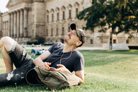 A young guy with a backpack or a student lies on the grass and listens to music or a podcast and enjoys.の写真素材