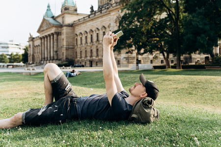A young guy with a backpack or a student is lying on the grass during a break or is resting and using a tablet. He is watching a video or reading or something else.の写真素材