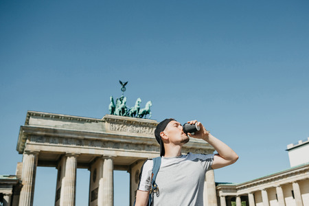 A person drinks from a disposable cup of coffee or another drink during a walk or sightseeing in Berlin in Germany. Ahead is the Brandenburg Gate.の写真素材