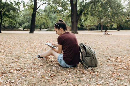 A young girl with a backpack sits in the autumn park and uses the tablet. She watches a video or reads or online or something else.の写真素材