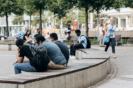 Germany, Leipzig, September 6, 2018: A group of students sits on a bench in a city park and communicates. In the background an African student is listening to music. Everyday city life.のeditorial素材
