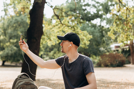A young guy or student talking on a cell phone via video connection or listening to music or watching a video in an autumn park.の写真素材
