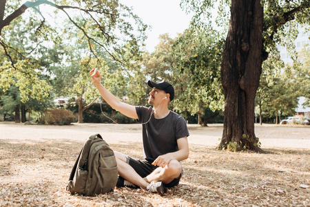 A young guy or student talking on a cell phone via video connection or listening to music or watching a video in an autumn park.の写真素材