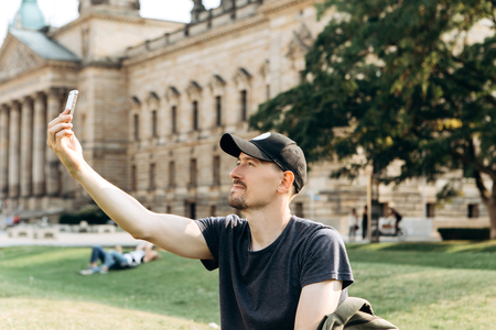 A young male tourist or student sits on the grass outside in Leipzig, Germany, talking on video or using a mobile phone to chat or take pictures.の写真素材