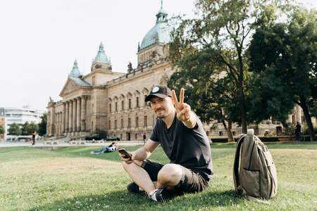 Young man tourist or student sitting on the grass in the streets of Leipzig in Germany and shows the hand gesture means peace.の写真素材