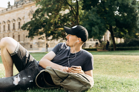A young guy with a backpack or a student lies on the grass and listens to music or a podcast and enjoys.の写真素材