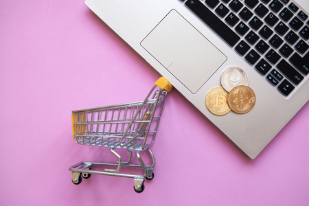 Conceptual photo of e-commerce using cryptocurrency. Computer, shopping trolley and Bitcoin coins or cryptocurrency on a trendy pink background.の写真素材