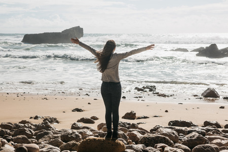 Rear view of a girl admiring the beautiful view of the Atlantic Ocean and the landscape and raising her hands up showing how she likes and how pleased she is.の写真素材