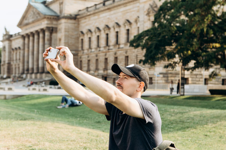 A young male tourist or student sits on the grass outside in Leipzig, Germany, talking on video or using a mobile phone to chat or take pictures.の写真素材