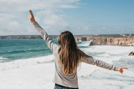 Rear view of a girl admiring the beautiful view of the Atlantic Ocean and the landscape and raising her hands up showing how pleased she is.の写真素材