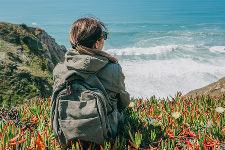A tourist girl with a backpack sits in solitude among flowers and plants on a hill and enjoys a...