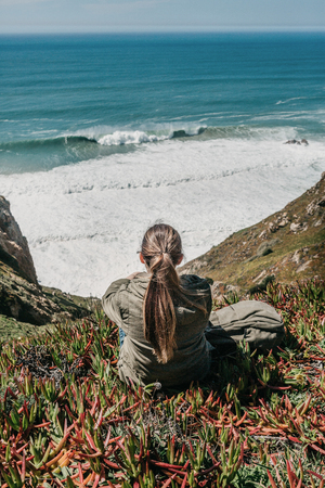 A tourist girl with a backpack sits in solitude among flowers and plants on a hill and enjoys a beautiful view of the Atlantic Ocean on a spring day.の写真素材