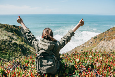 A girl tourist with a backpack sits on a hill, enjoys a beautiful view of the Atlantic Ocean and raises hands showing how pleased she is.の写真素材