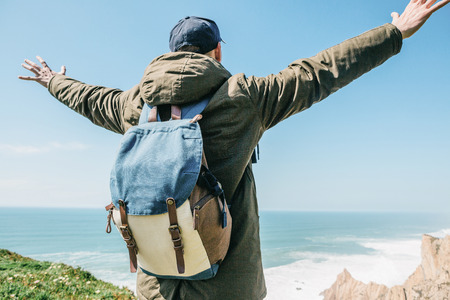 A tourist with a backpack on a cliff against the background of the Atlantic Ocean raised his hands and shows how he is free and happy.の写真素材
