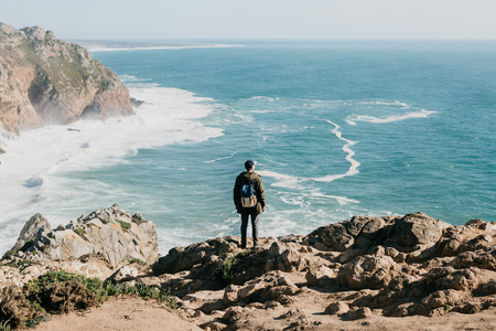 A man tourist with a backpack stands in solitude at Cape Roca in Portugal and enjoys a beautiful view of the Atlantic Ocean.の写真素材