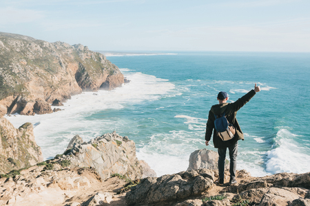 A tourist with a backpack on a cliff against the background of the Atlantic Ocean raised his hand and shows how he is free and happy.の写真素材