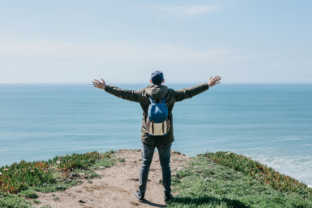 A tourist with a backpack on a cliff against the background of the Atlantic Ocean raised his hands and shows how he is free and happy.の写真素材