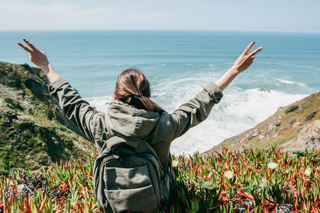 A girl tourist with a backpack sits on a hill, enjoys a beautiful view of the Atlantic Ocean and raises hands showing how pleased she is.の写真素材