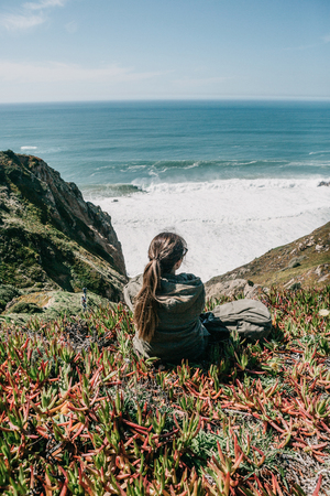A tourist girl with a backpack sits in solitude among flowers and plants on a hill and enjoys a beautiful view of the Atlantic Ocean on a spring day.の写真素材