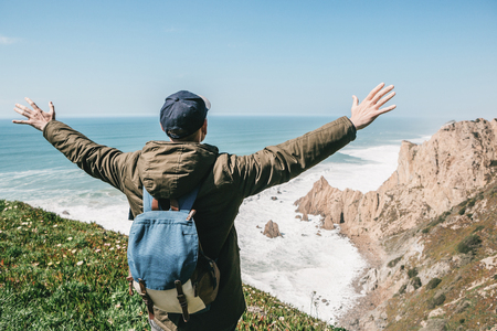A tourist with a backpack on a cliff against the background of the Atlantic Ocean raised his hands and shows how he is free and happy.の写真素材