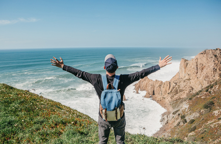 A tourist with a backpack on a cliff against the background of the Atlantic Ocean raised his hands and shows how he is free and happy.の写真素材