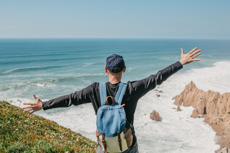 A tourist with a backpack on a cliff against the background of the Atlantic Ocean raised his hands and shows how he is free and happy.の写真素材