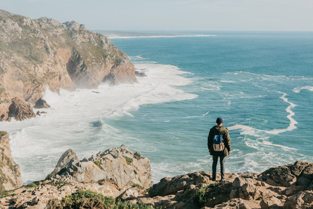 A man tourist with a backpack stands in solitude at Cape Roca in Portugal and enjoys a beautiful view of the Atlantic Ocean.の写真素材