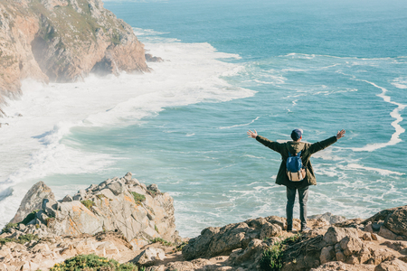 A tourist with a backpack on a cliff against the background of the Atlantic Ocean raised his hands and shows how he is free and happy.の写真素材