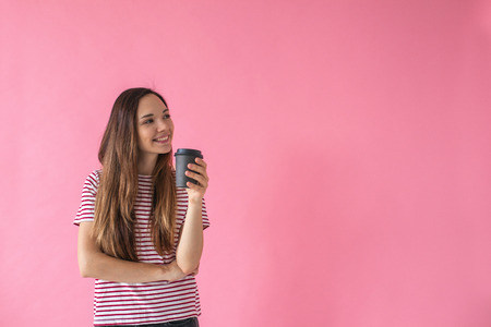 Beautiful smiling positive girl drinks coffee on a pink background. Coffee break.の写真素材