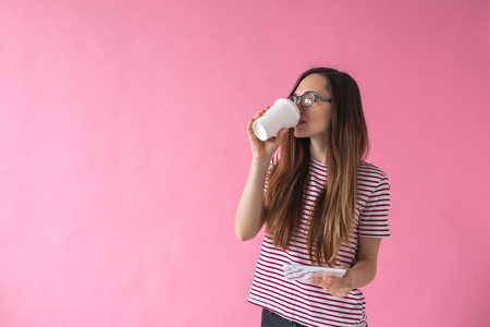 A beautiful young girl drinks coffee or another drink and uses the tablet on a pink background.の写真素材