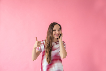 Positive beautiful young girl uses a cell phone and shows thumb up sign meaning OK.の写真素材