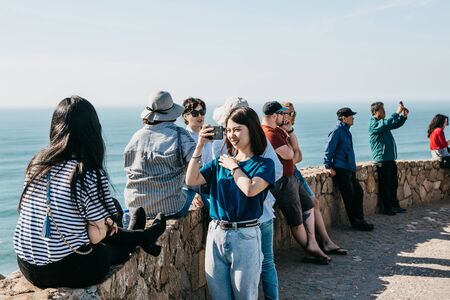 Portugal, Sintra, June 26, 2018: Asian tourist girl takes a picture of her friend on a cell phone at Cape Roca. Nearby are other tourists.のeditorial素材