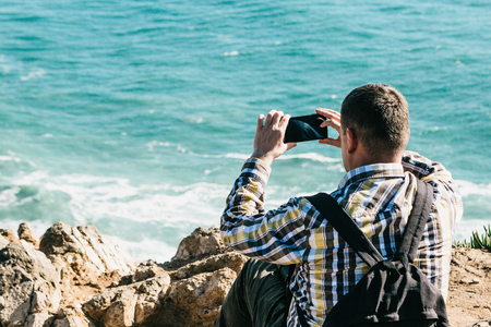 An adult unrecognizable male backpacker photographs on a cell phone a beautiful view of the Atlantic Ocean from the shore.の写真素材