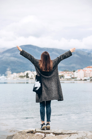A trendy girl stands off the coast of the sea and looks into the distance and raises her hands up showing how happy she is.の写真素材